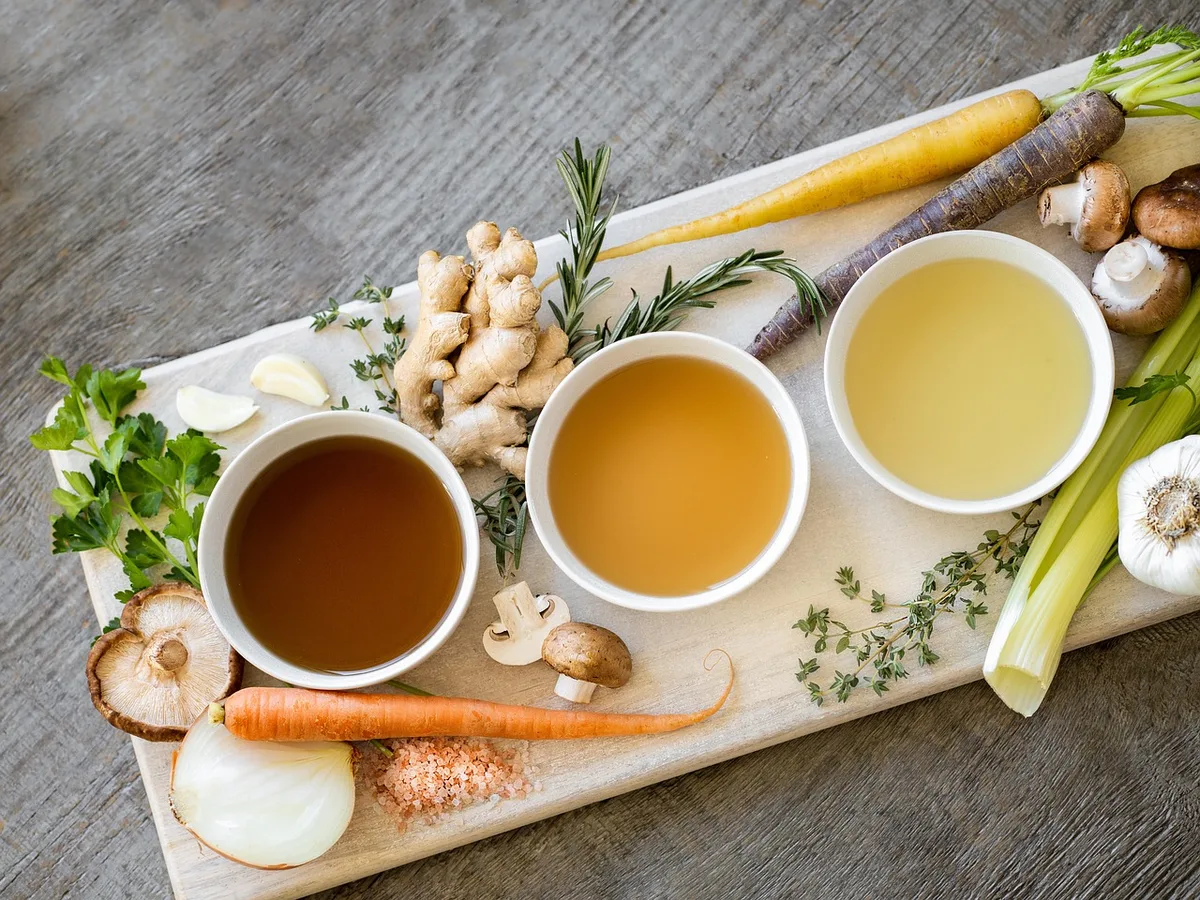 Three bowls of broth in different shades, surrounded by fresh ginger, mushrooms, carrots, garlic, celery and herbs