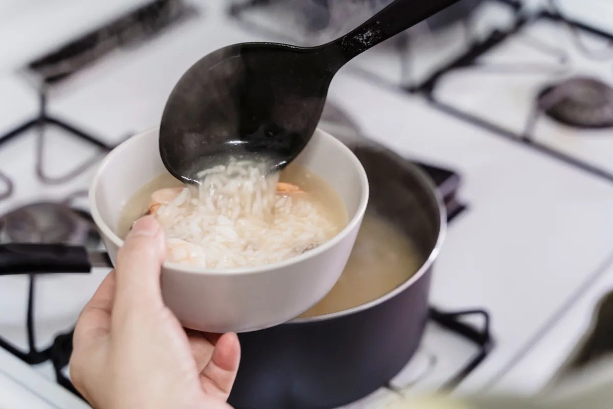 Plain rice porridge being ladled from a pot on the stove into a white bowl