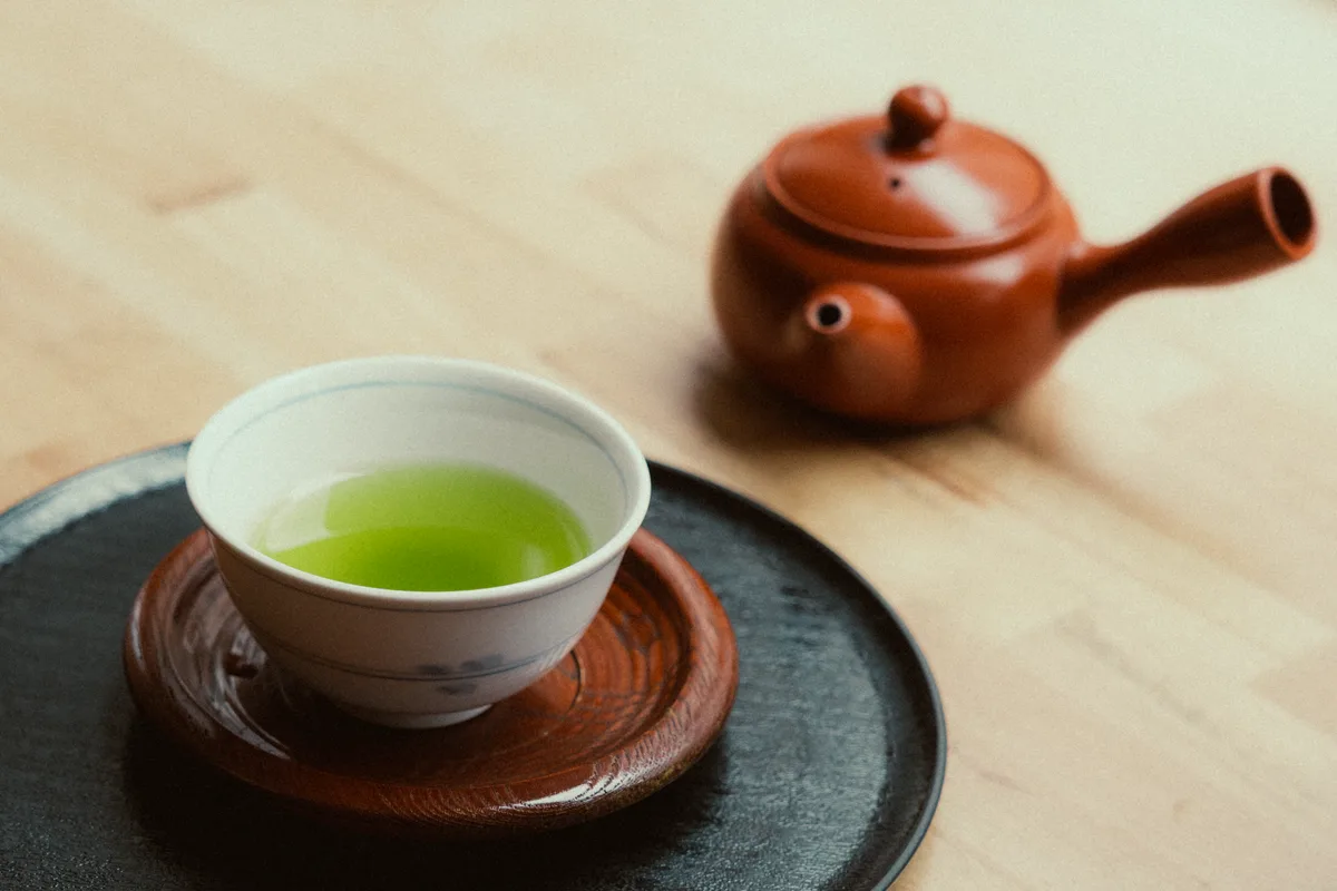 A small cup of bright green tea on a wooden saucer, with a clay teapot behind it