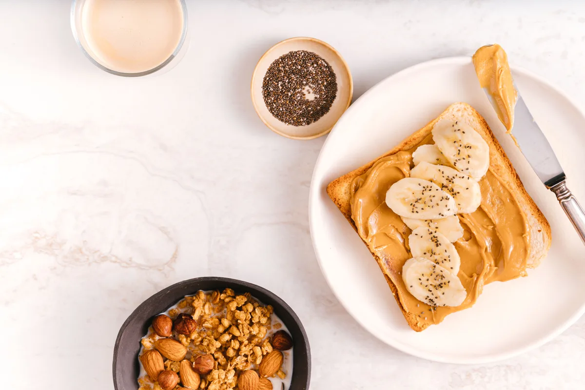 Toast spread with nut butter and topped with banana slices and chia seeds, next to a small bowl of mixed nuts