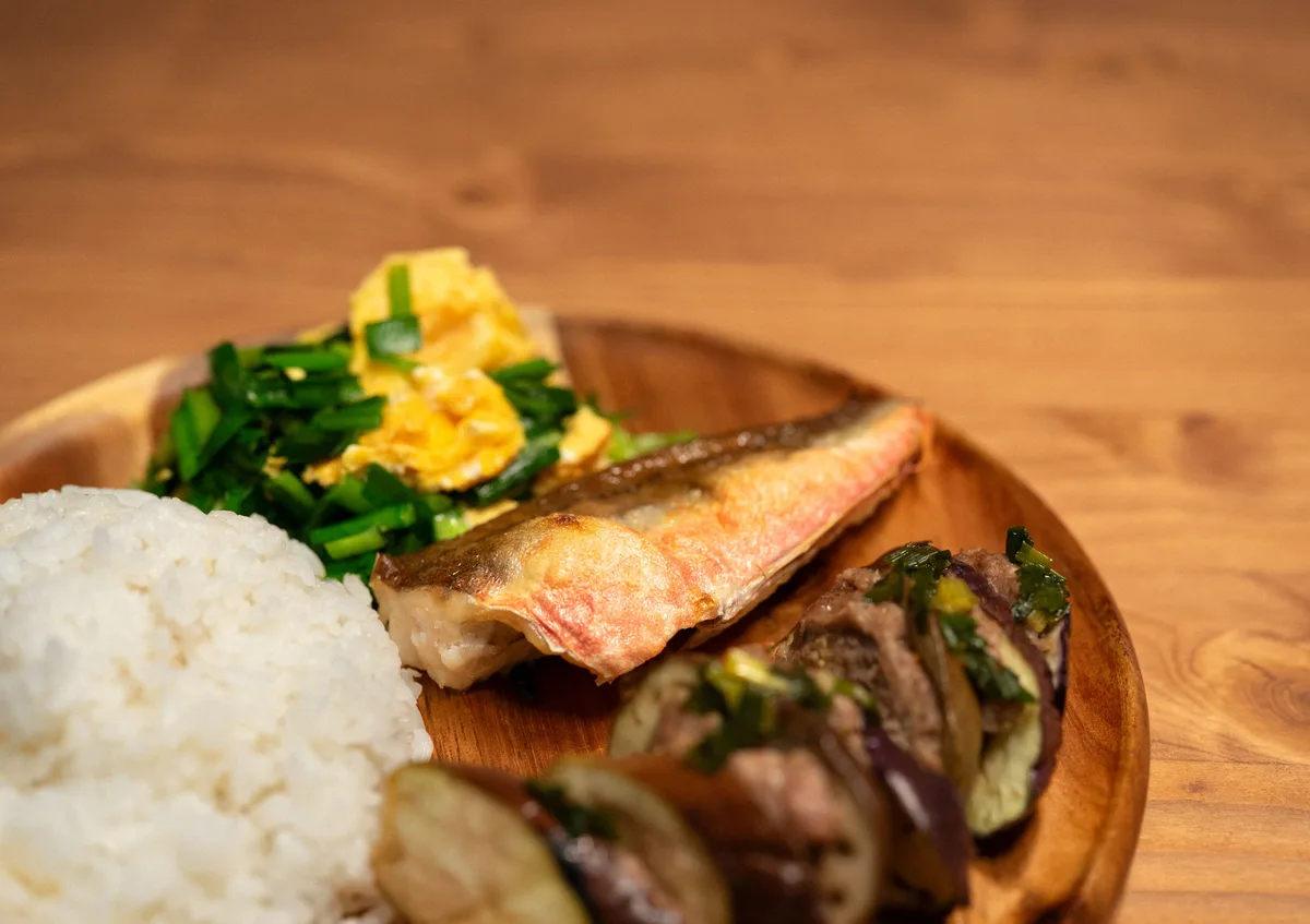 A wooden plate with grilled fish, white rice, scrambled egg with chives and roasted eggplant