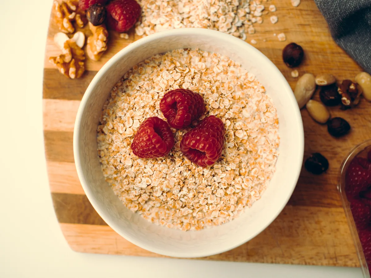 A bowl of oats topped with raspberries, with walnuts and almonds on a wooden board