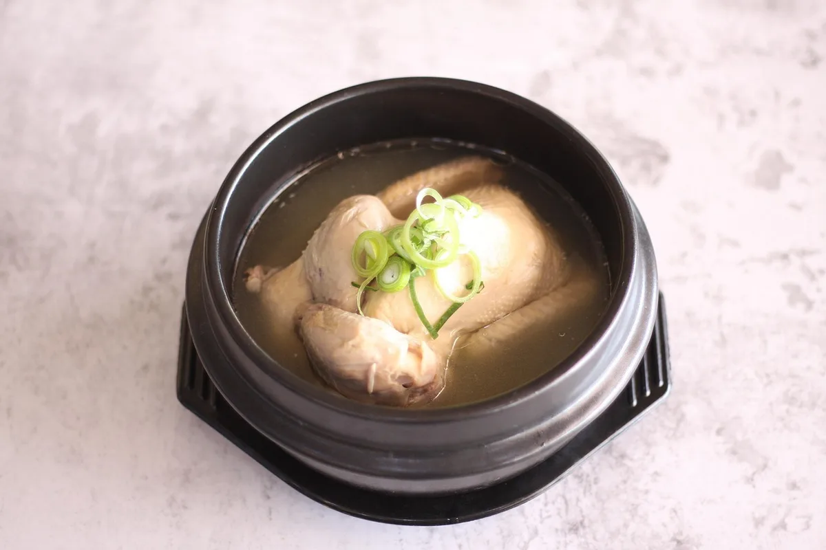 Samgyetang, a whole small chicken in clear broth topped with sliced green onion, served in a black stone pot