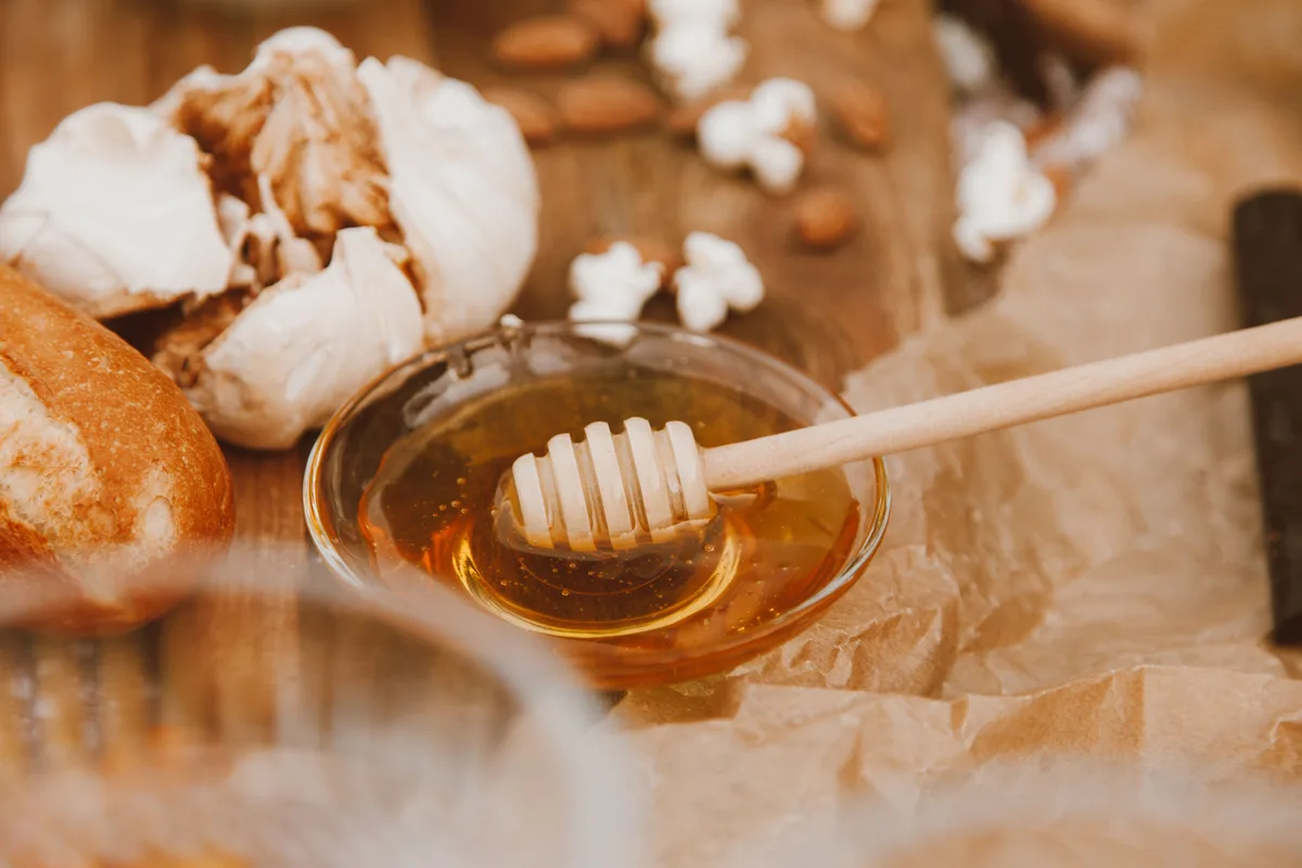 Garlic bulbs next to a small glass bowl of honey with a wooden honey dipper