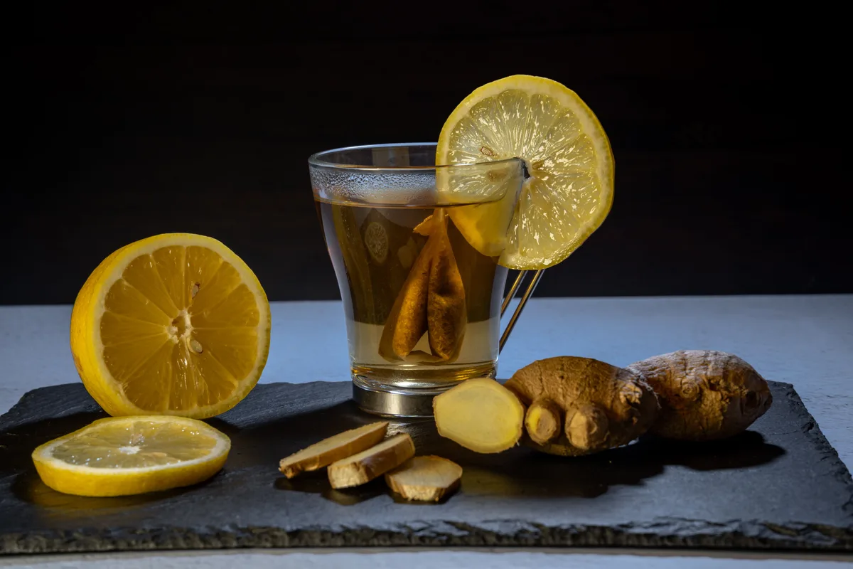 A glass of ginger lemon tea with a lemon slice on the rim, fresh ginger root and lemon halves on a slate board
