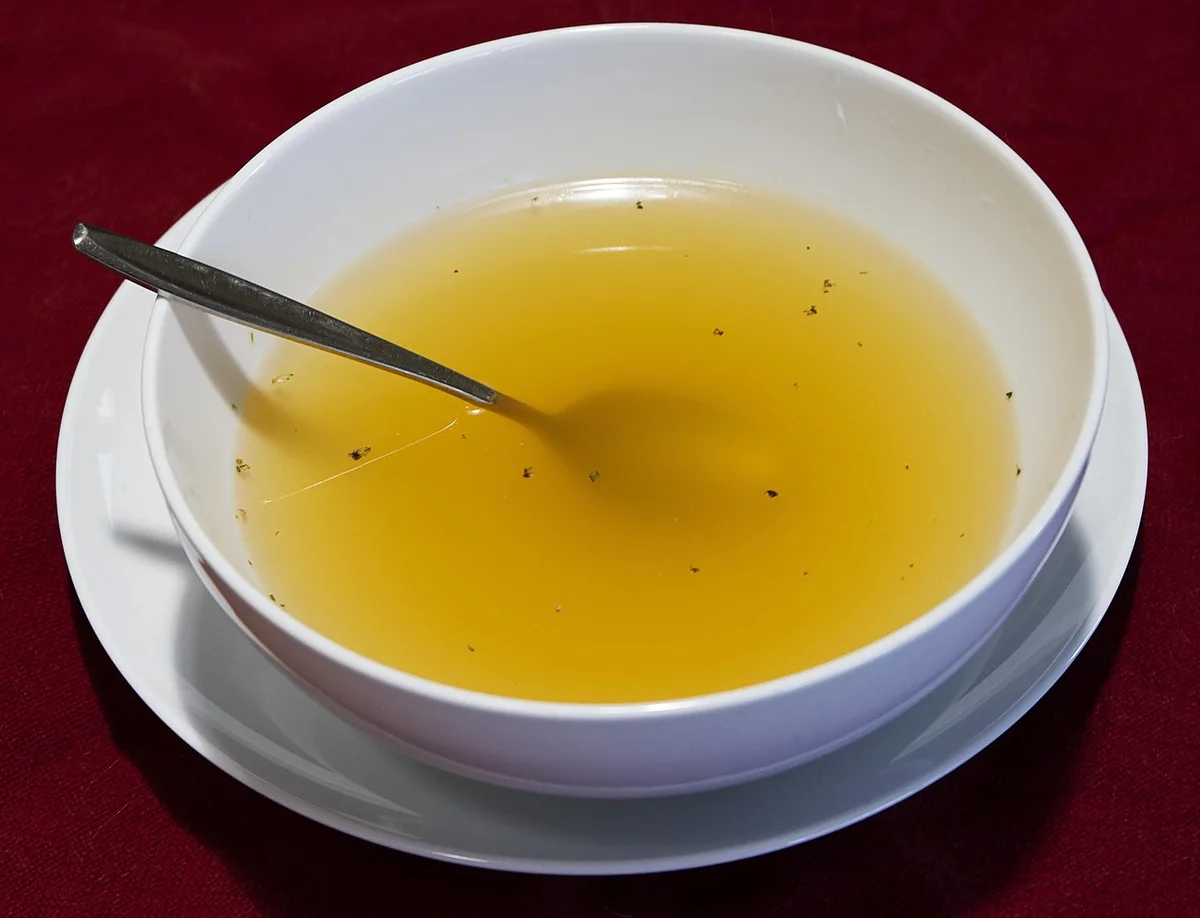 A white bowl of clear golden broth with a spoon resting in it