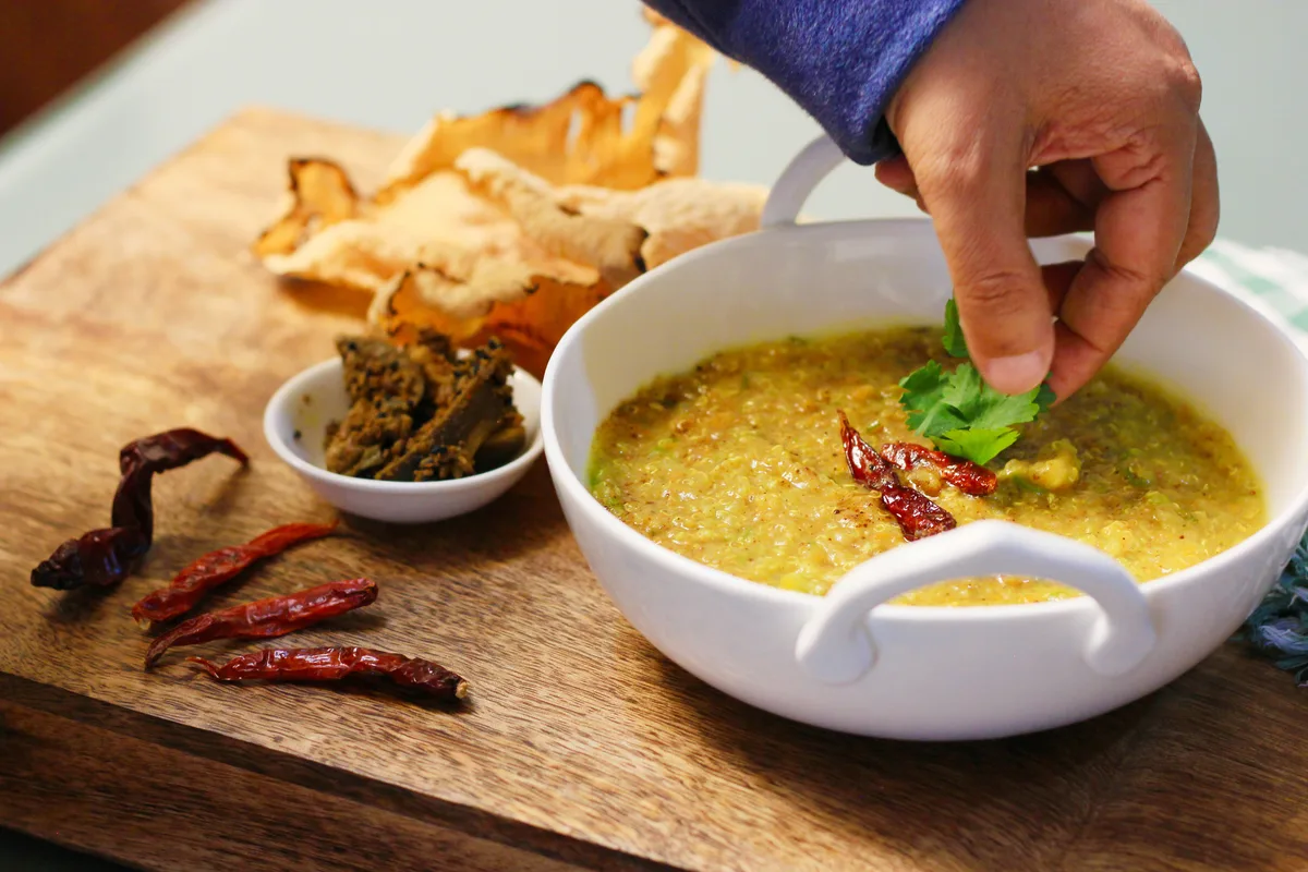 A hand adding coriander leaves to a bowl of yellow khichdi, with dried red chillies and papadum on the side