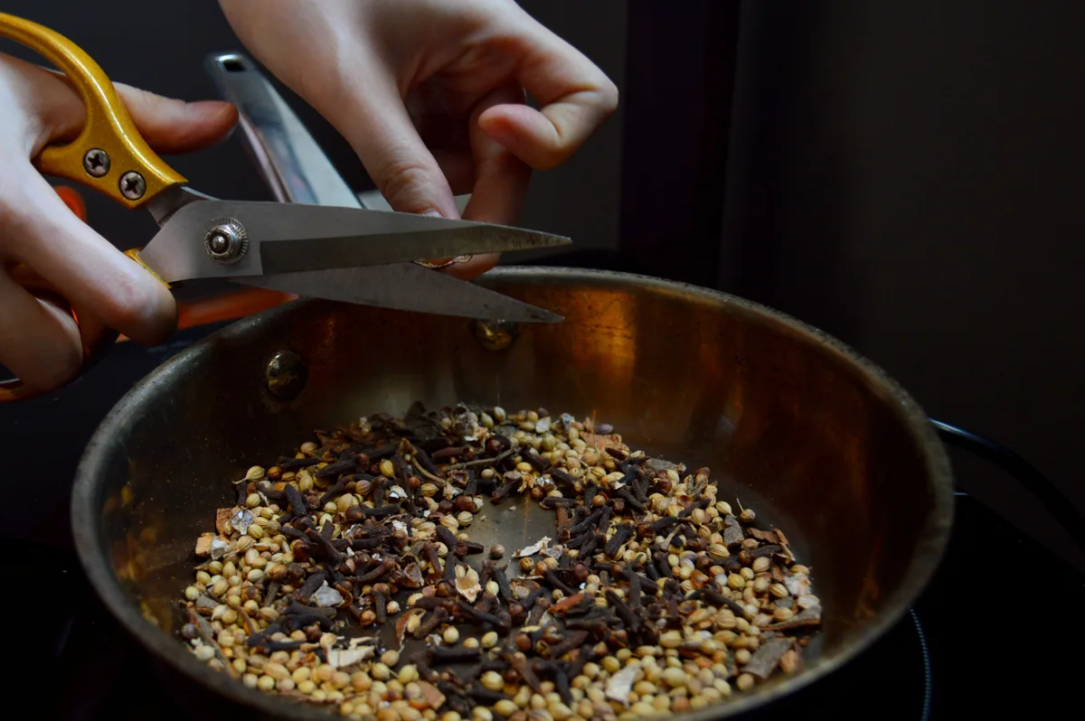 Coriander seeds and cloves toasting in a copper pan while a cook works over it with kitchen scissors