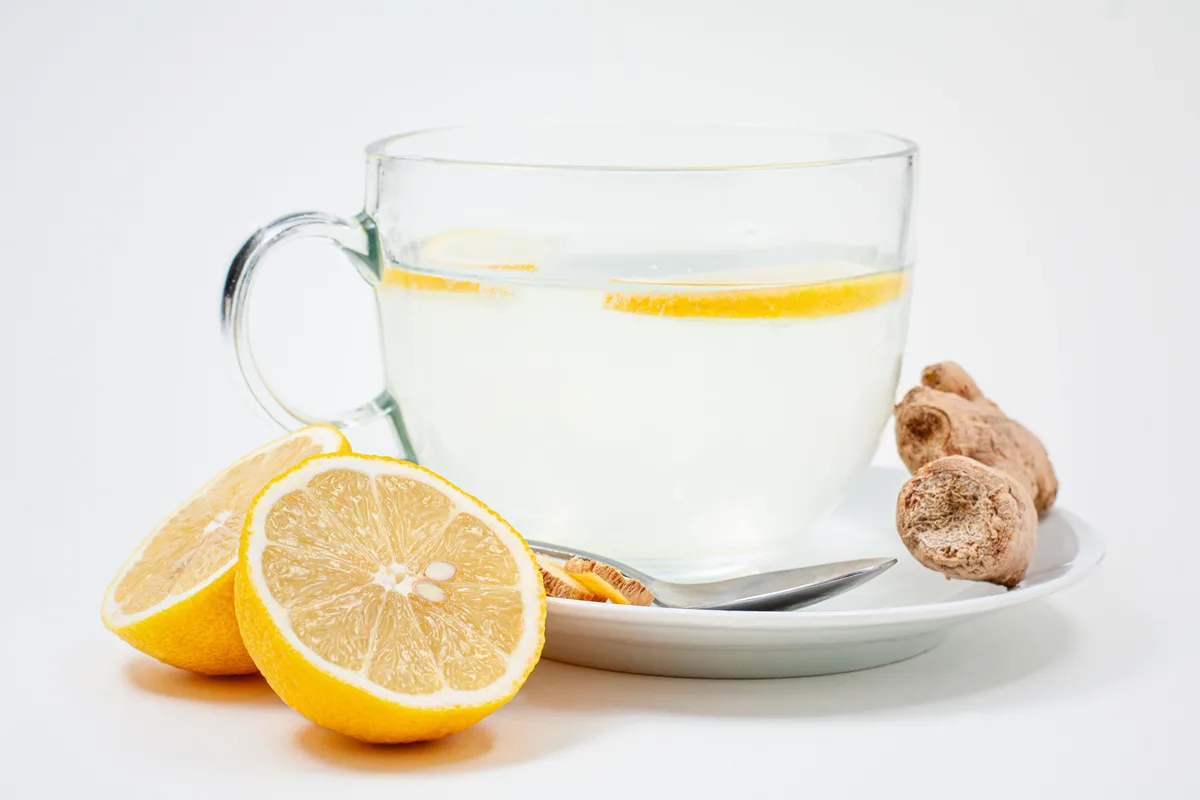 A glass cup of warm water with lemon slices, next to a halved lemon and a piece of fresh ginger