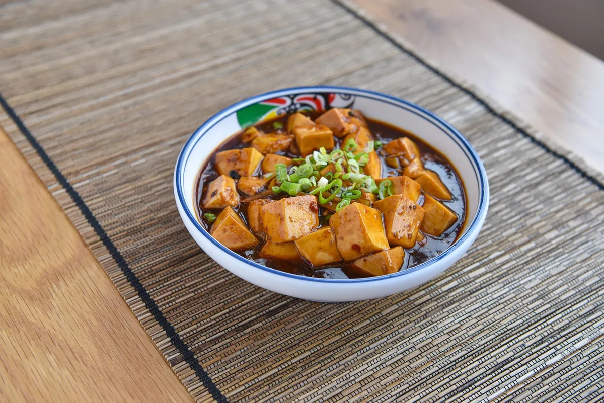 Mapo tofu in a blue and white bowl, cubes of tofu in dark chilli sauce topped with sliced scallions