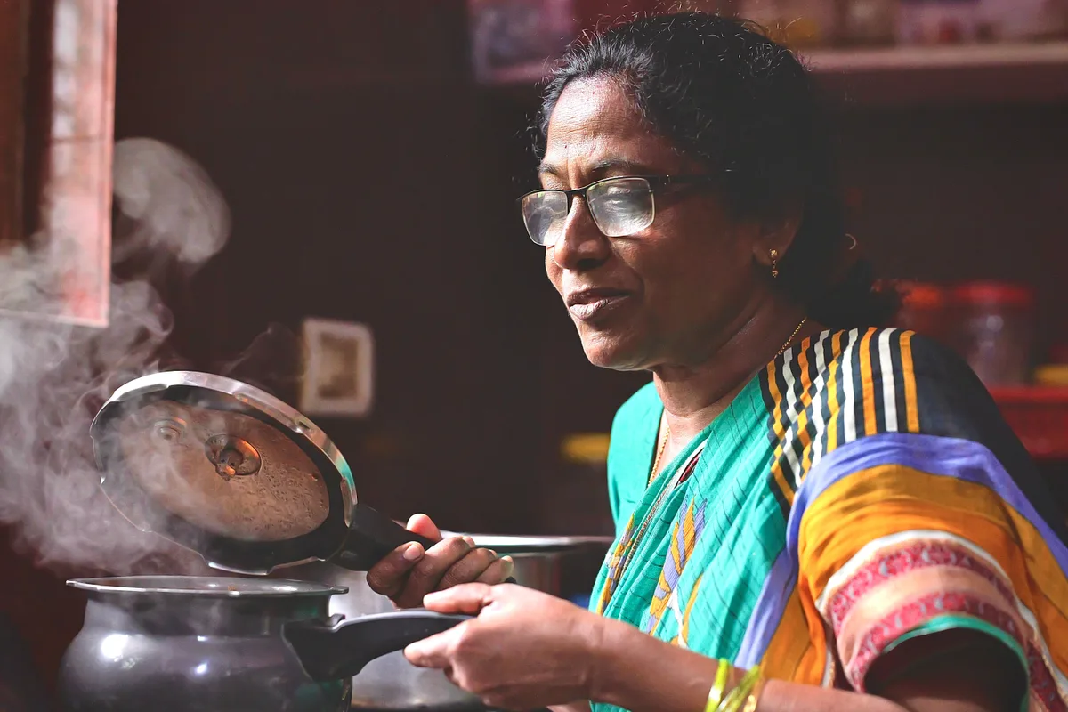 A woman lifts the lid off a steaming pressure cooker in her kitchen