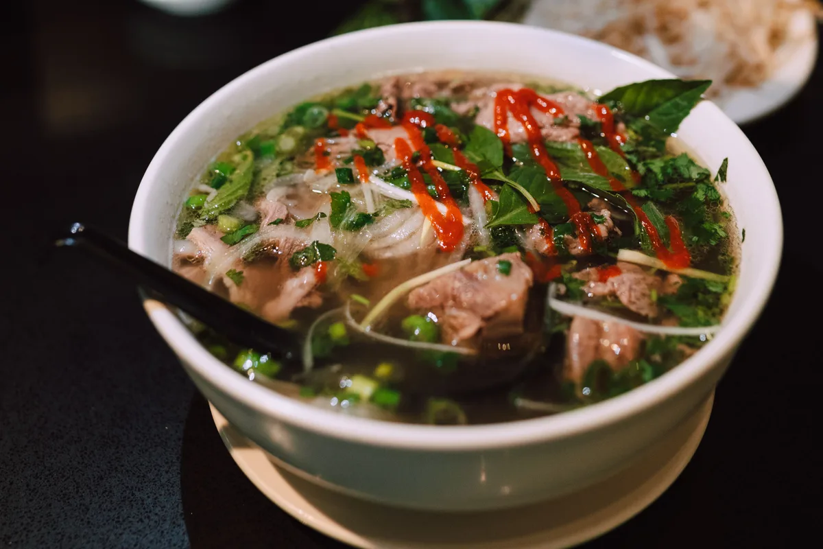 A bowl of beef pho with herbs and a drizzle of chilli sauce on a dark table