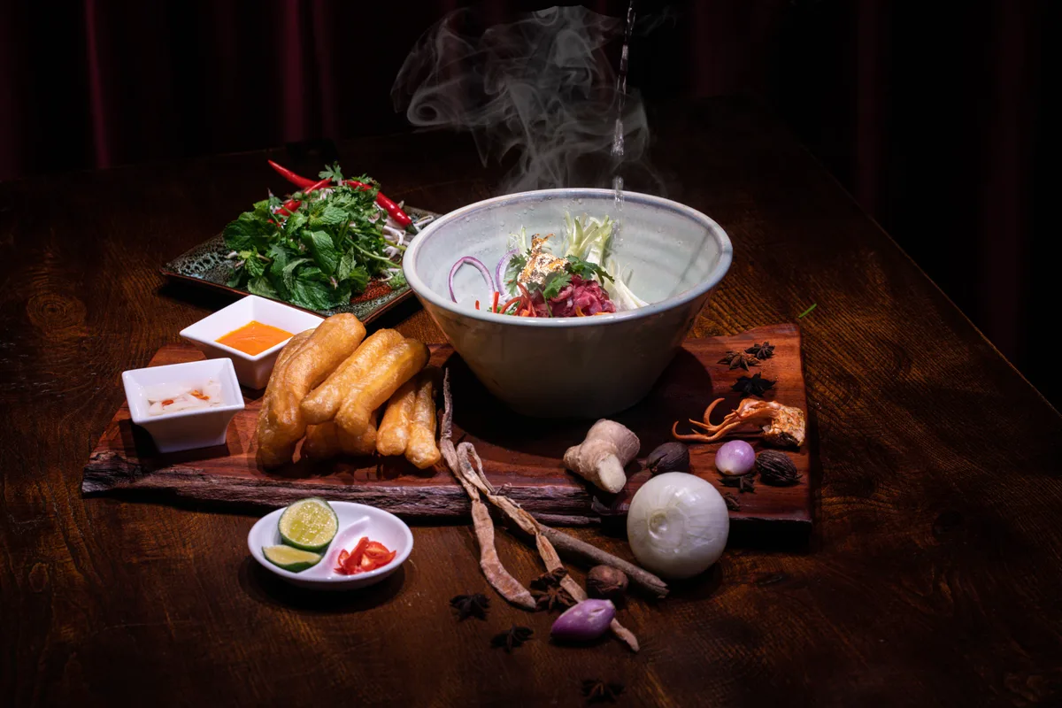 A steaming bowl of pho on a dark table with fried dough sticks, fresh herbs, lime, ginger, shallots, star anise and cinnamon bark