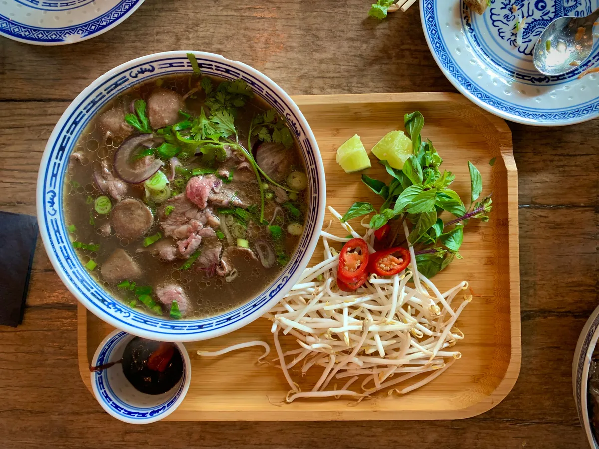 Pho with sliced beef and meatballs beside a tray of bean sprouts, basil, lime and red chillies